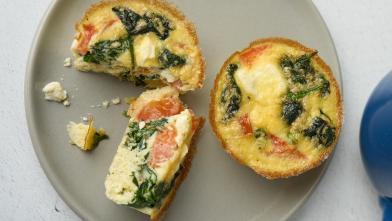 Overhead view of three small spinach and tomato quiche tarts on a gray plate next to a blue bowl.