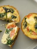 Overhead view of three small spinach and tomato quiche tarts on a gray plate next to a blue bowl.