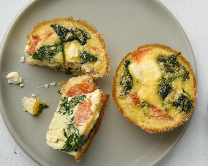 Overhead view of three small spinach and tomato quiche tarts on a gray plate next to a blue bowl.