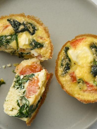 Overhead view of three small spinach and tomato quiche tarts on a gray plate next to a blue bowl.