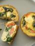 Overhead view of three small spinach and tomato quiche tarts on a gray plate next to a blue bowl.