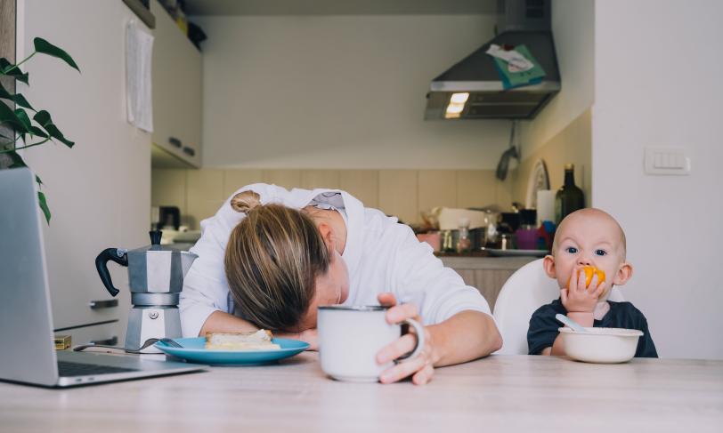 Tired Woman next to Child eating an orange at a table in the kitchen.