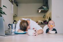 Tired Woman next to Child eating an orange at a table in the kitchen.