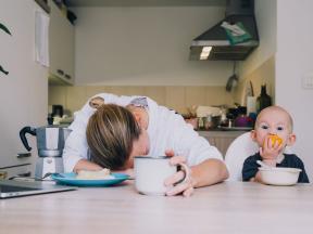 Tired Woman next to Child eating an orange at a table in the kitchen.