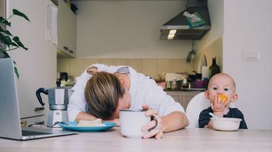 Tired Woman next to Child eating an orange at a table in the kitchen.